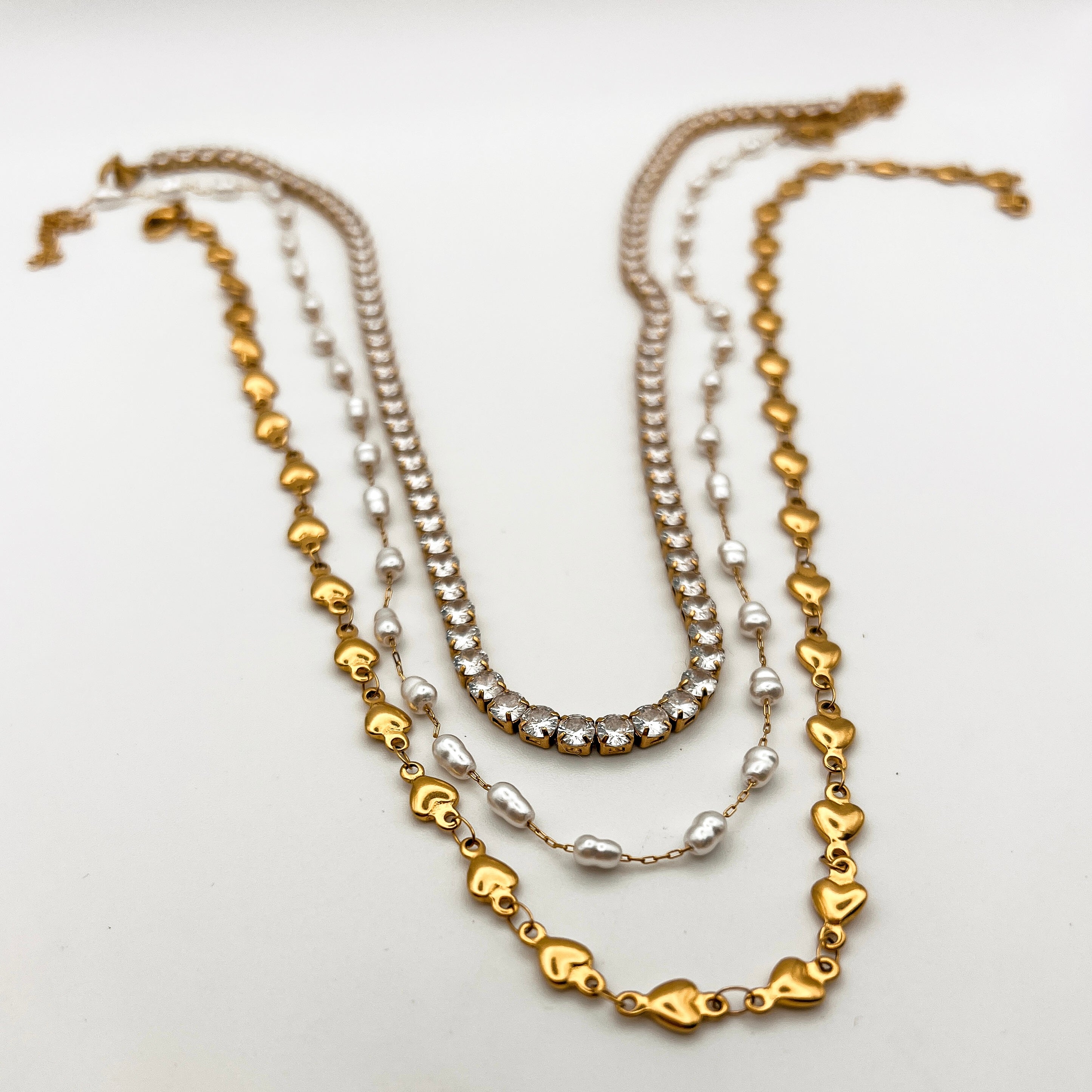 A tennis necklace, a baroque pearl necklace, and a gold heart chain neckalce laying on a white surface